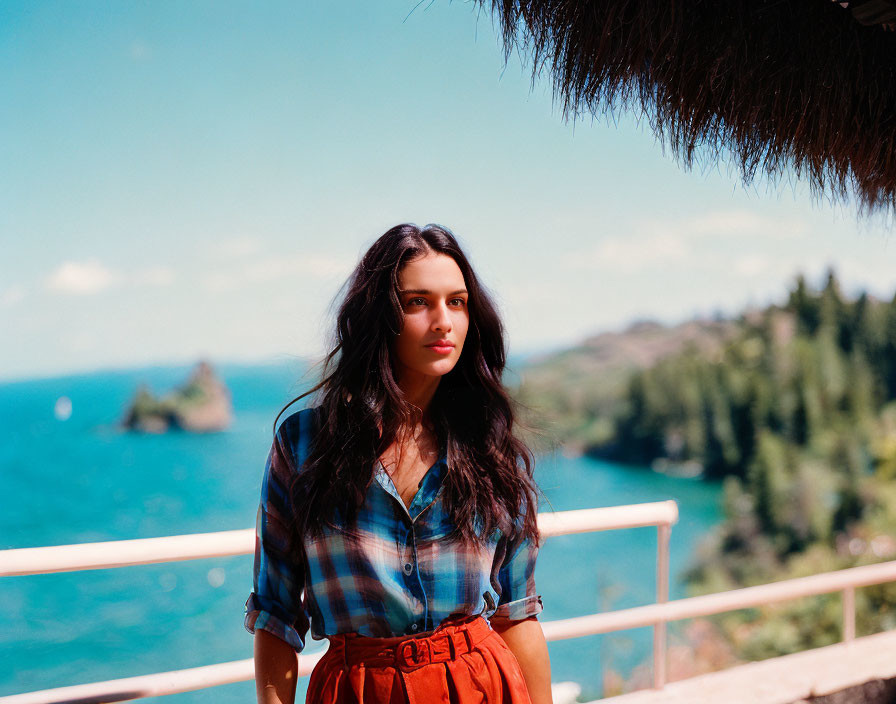 Woman in plaid shirt and red skirt on balcony with thatched roof, scenic landscape view