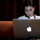 Focused woman working on MacBook in dimly lit room with curtains and orange chair.