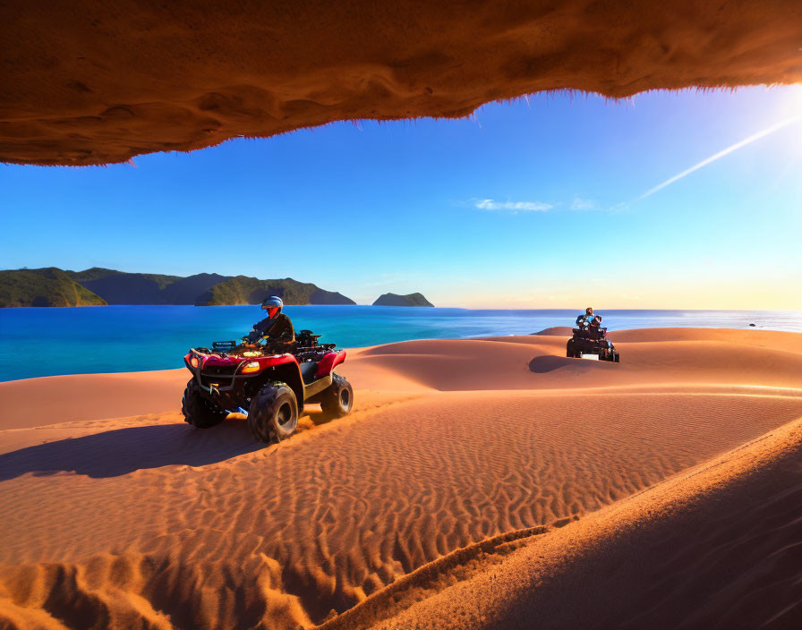 Two people riding ATVs on sandy dunes near ocean with rock formation and clear blue sky.