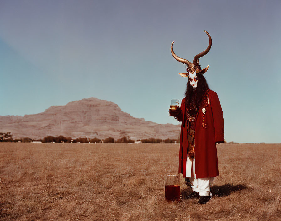 Person in red coat and white pants with horned mask holding cup in field with mountain backdrop.