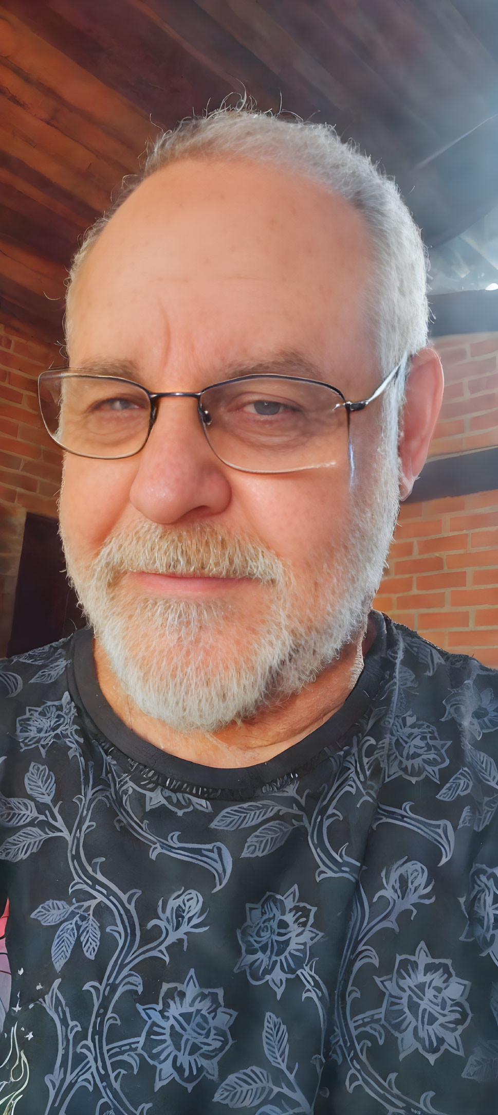 Smiling man with grey beard and glasses in black shirt with floral pattern by brick structure