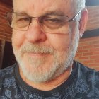 Smiling man with grey beard and glasses in black shirt with floral pattern by brick structure