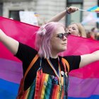 Pink-haired person with flower accessory holding bi-pride flag at outdoor event.