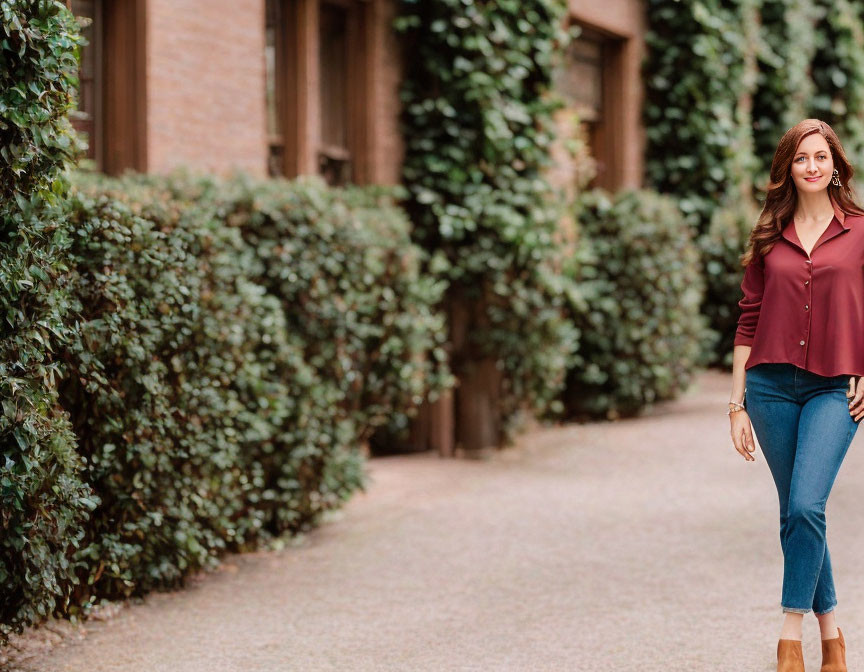 Smiling woman in burgundy blouse walking by green hedge and brick building