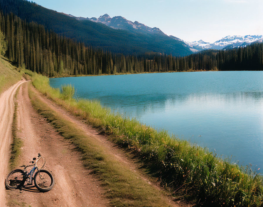 Mountain bike by dirt track at serene lake with forested hills & snowy mountains