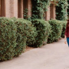 Smiling woman in burgundy blouse walking by green hedge and brick building