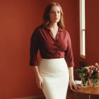 Woman in burgundy blouse and cream skirt by window with flowers against terracotta wall