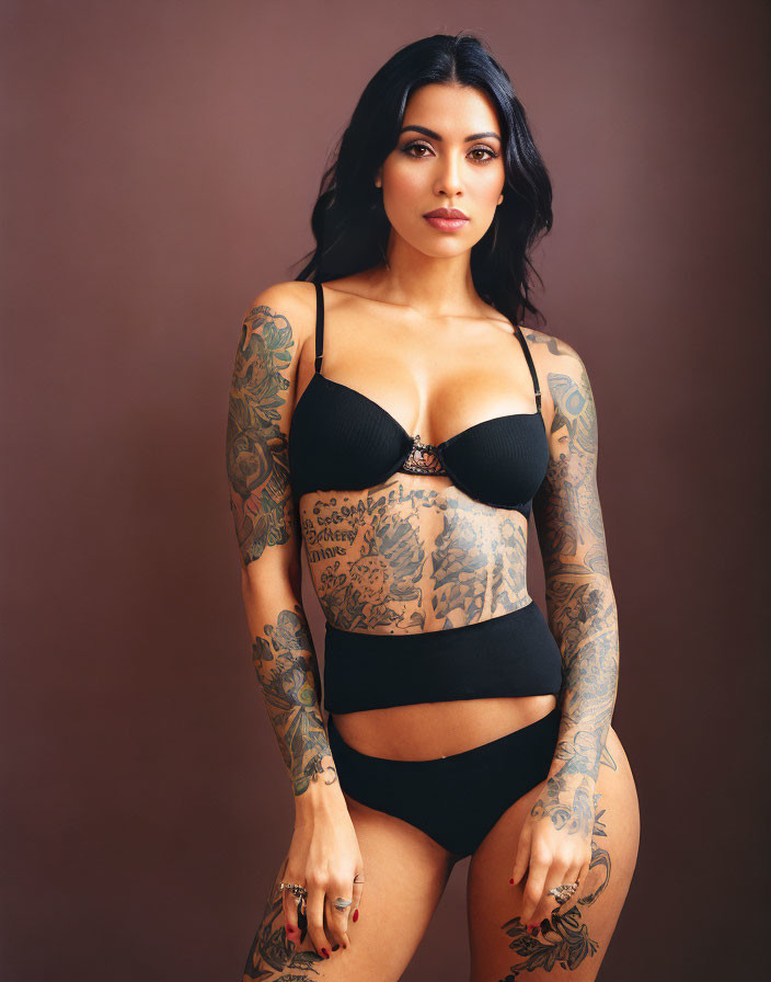 Woman with full-sleeve tattoos in black lingerie and high-waist bottom on brown backdrop