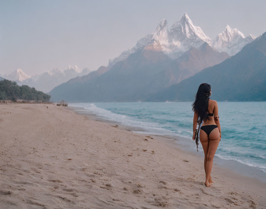 Bikini-clad person on sandy beach admires snow-capped mountains