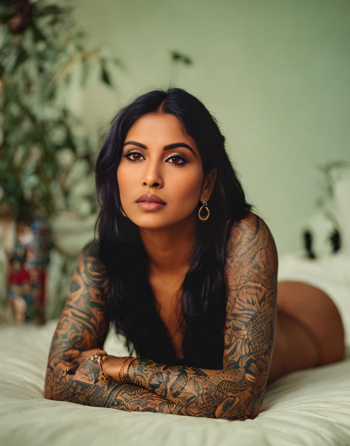 Dark-haired woman with sleeve tattoos against green backdrop and potted plants, adorned with gold jewelry