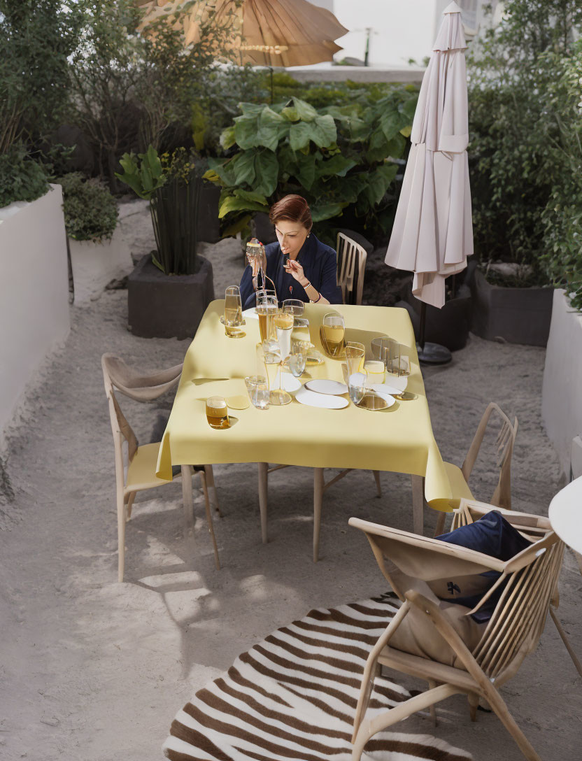 Solitary figure at elegantly set outdoor table with yellow tablecloth, surrounded by plants and umbrella.