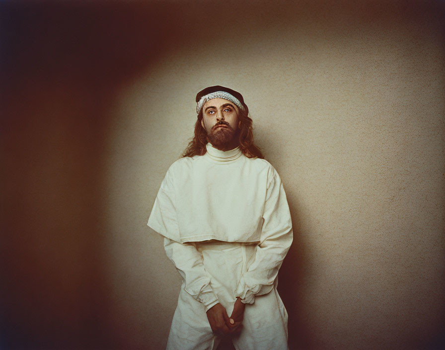 Bearded man in white turtleneck and keffiyeh, looking pensive against plain backdrop