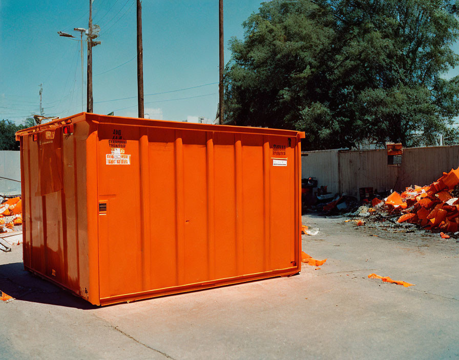 Orange dumpster in industrial area with scattered debris under clear blue sky