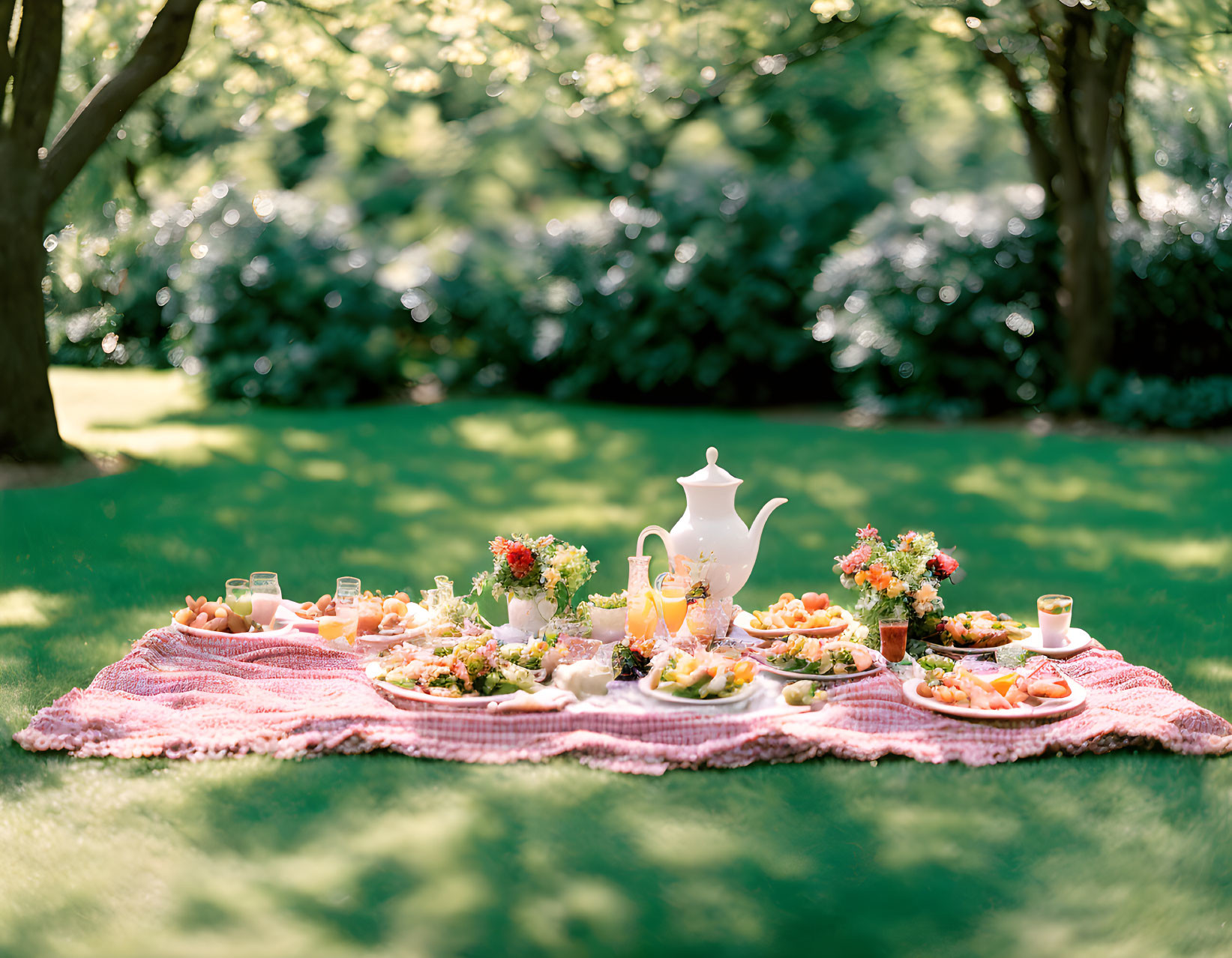 Picnic setup with tea pot, glasses, plates on pink blanket in lush greenery