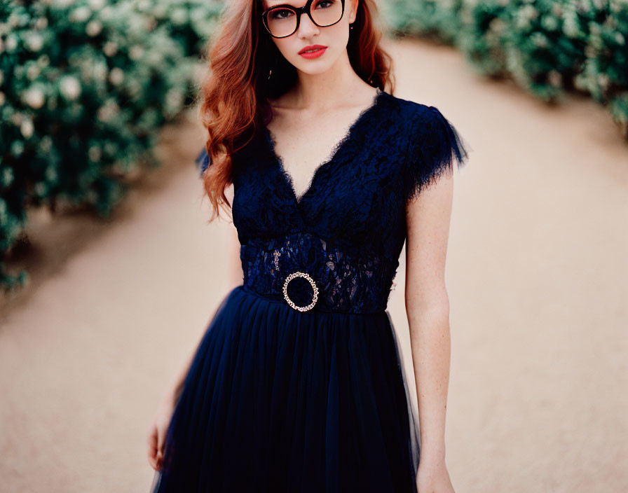 Red-haired woman in large glasses wearing blue lace dress with jeweled waistband amidst bushes