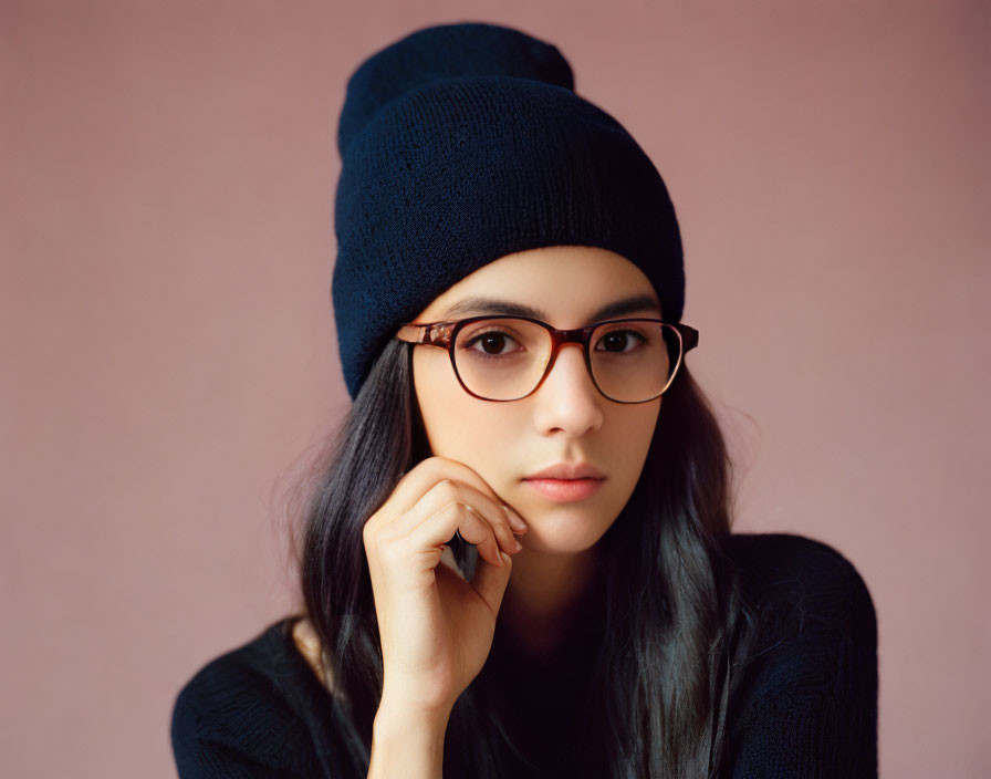 Long-haired person in glasses and beanie poses thoughtfully on pink background