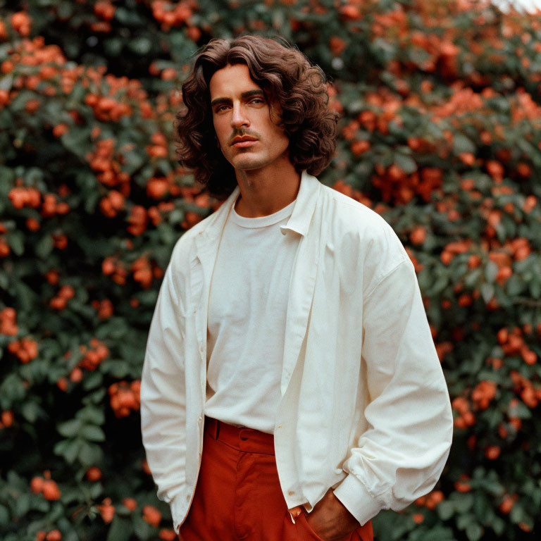 Man with Long Curly Hair in White Shirt and Red Pants by Bush with Orange Flowers