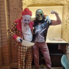 Two clowns in painted faces and theatrical outfits against rustic backdrop with yellow wall and arched doorway