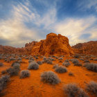 Sunset desert landscape with rock formations, sand dunes, and cloudy sky.