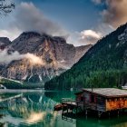 Snow-capped mountains reflected in serene lake with cabin on pier, pine trees, clear sunset sky