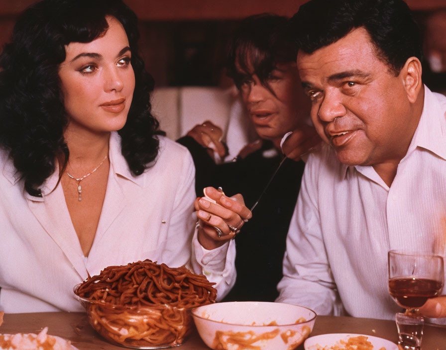 Man and woman at table with bowl of spaghetti and wine, dressed in white.