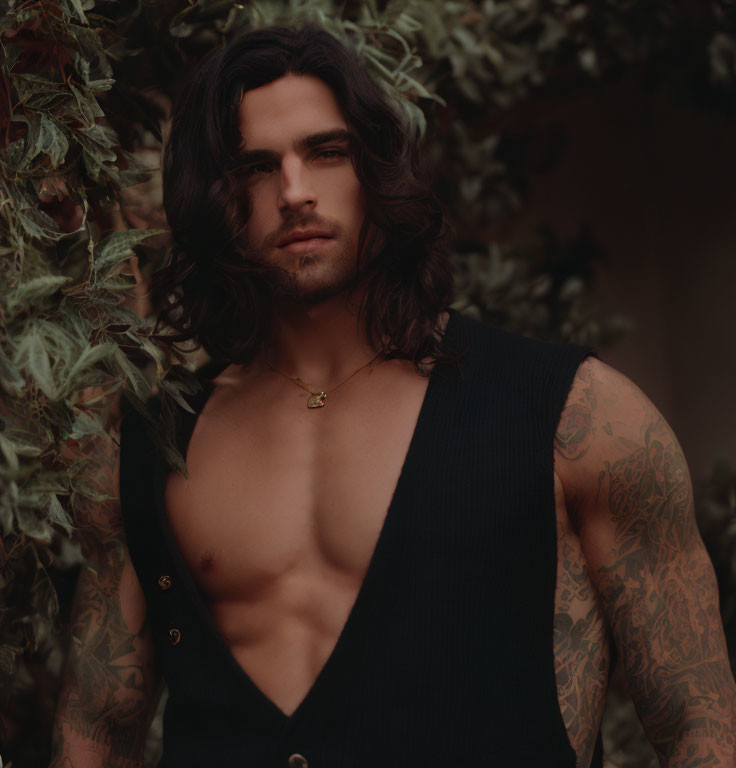 Man with Shoulder-Length Dark Hair and Tattoos in Sleeveless Black Top Among Leafy Greens