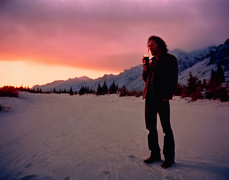 Person standing on snowy road at dusk with cup, vivid sunset skies, and silhouetted mountains