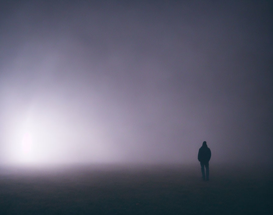 Mysterious lone figure walking towards bright light in misty fog