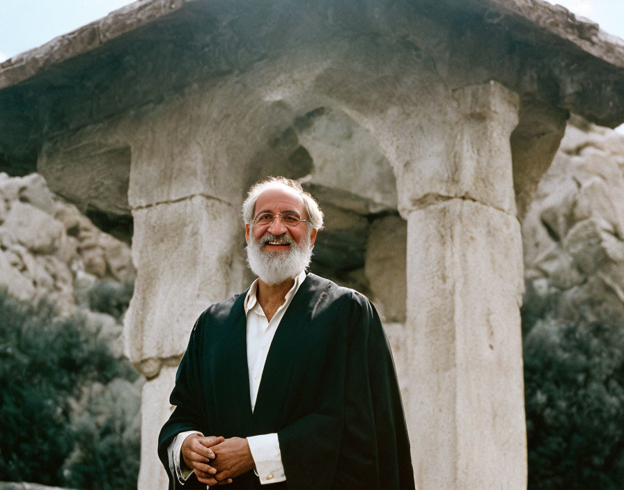 Elder man with white beard smiling near stone columns and greenery