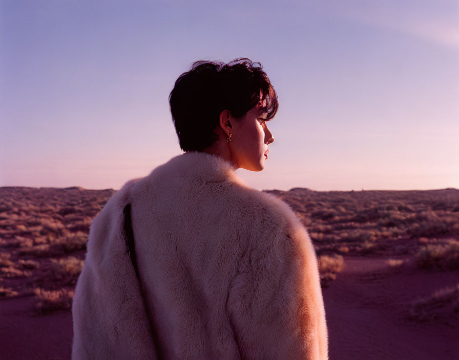 Person in White Fluffy Coat Facing Sunset on Sandy Landscape