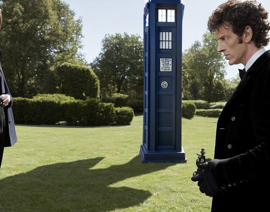 Man in suit with device next to blue police box on sunny lawn