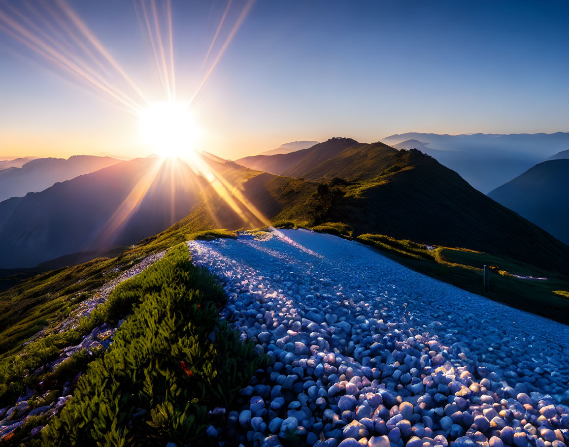 Mountain landscape at sunrise with vibrant blue sky and sun rays over rocky path.