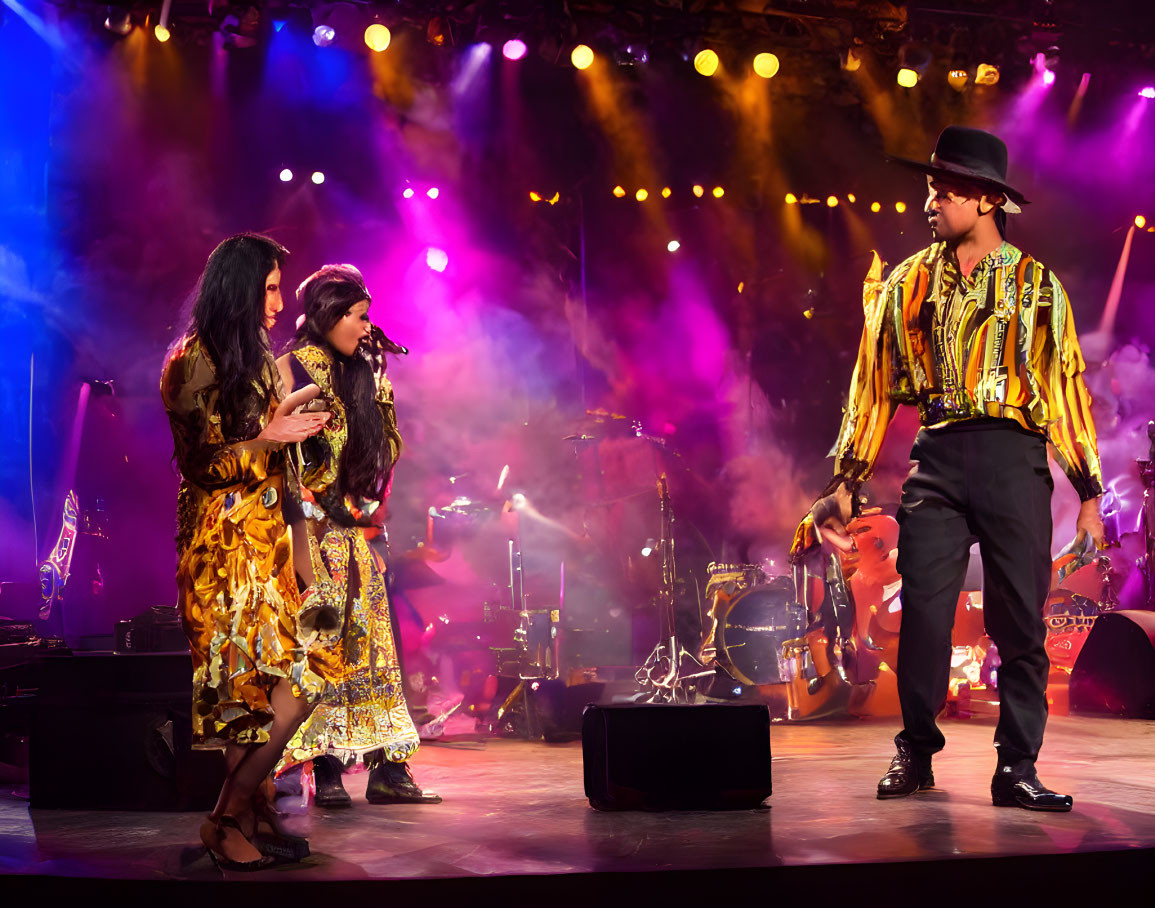 Three performers on stage with band setup; one in black hat and colorful jacket, others in patterned