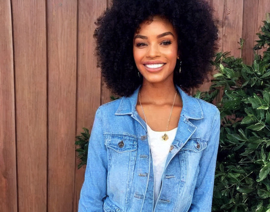 Smiling woman with curly hair in denim jacket by wooden wall