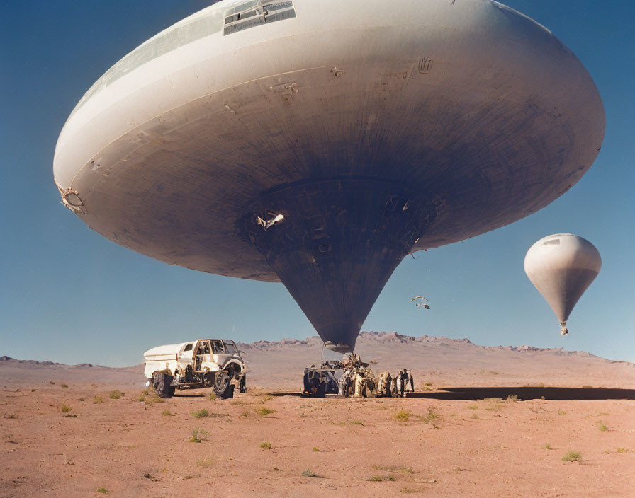 Large disc-shaped object hovers above desert scene with people and vehicles, smaller balloon in distance