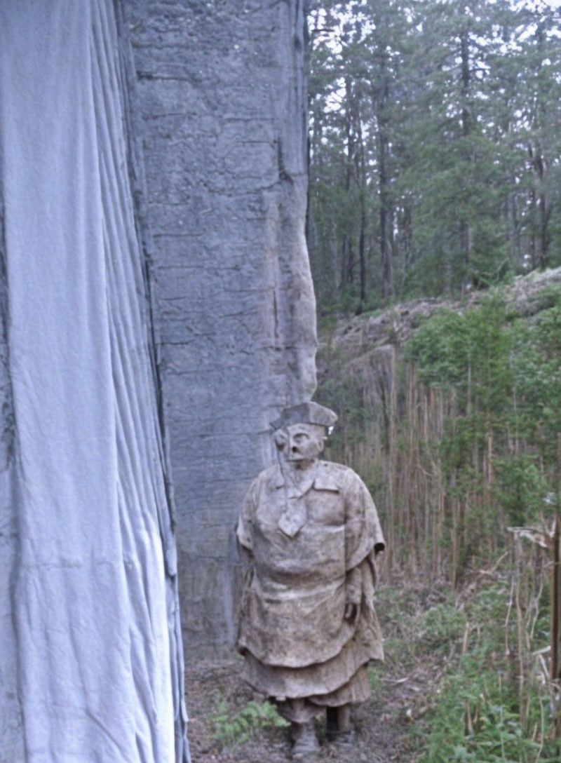 Stone sculpture of figure in cloak and hat blending with forest rock face