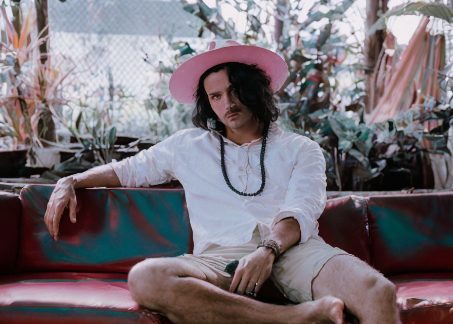 Man with shoulder-length hair in pink hat and white shirt sitting on colorful couch surrounded by plants.