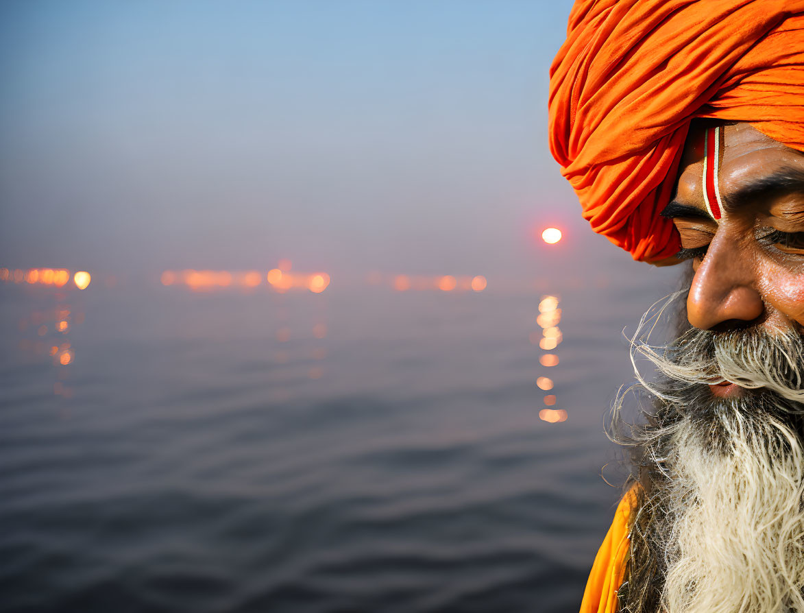 Profile of man in orange turban and white beard by serene water body at sunrise.