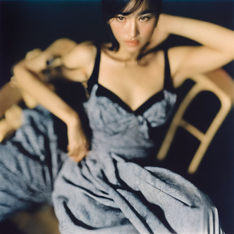 Woman in black top and blue skirt sitting on wooden chair gazes at camera