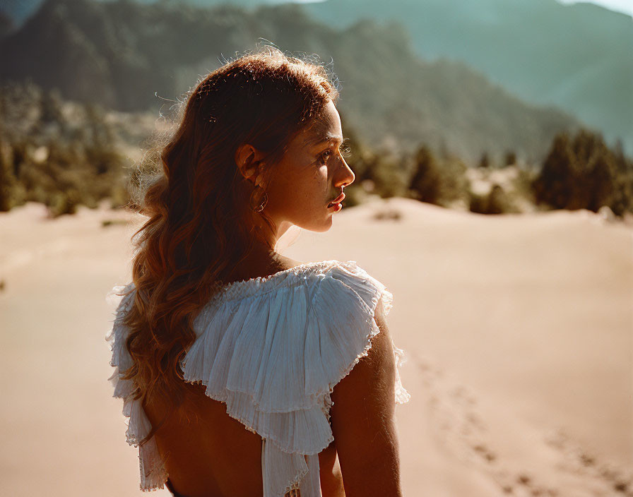 Woman in white ruffled dress gazes at mountains and sand dunes in warm sunlight