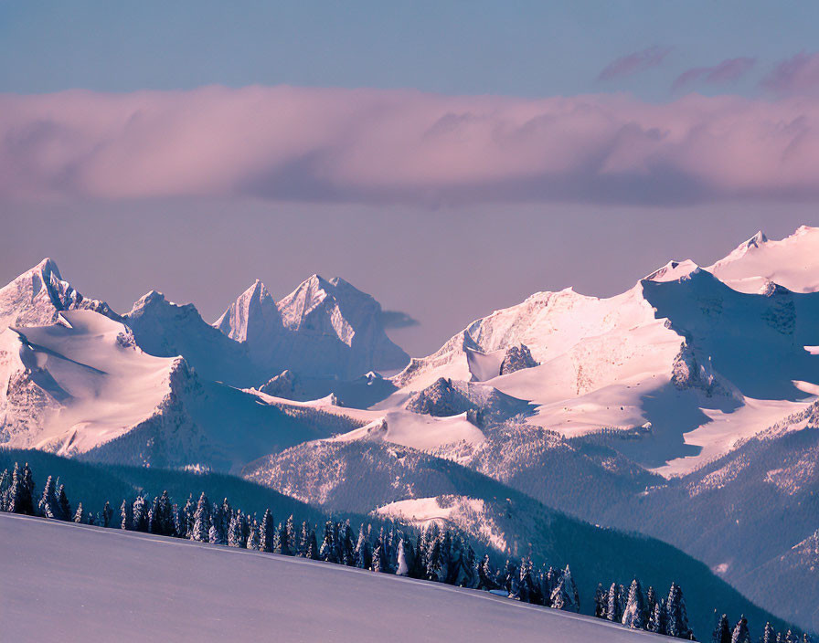 Snow-covered mountain peaks at sunset with tree-lined ridge.