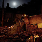 Nighttime campfire scene in forest clearing with tents, vehicles, and full moon.