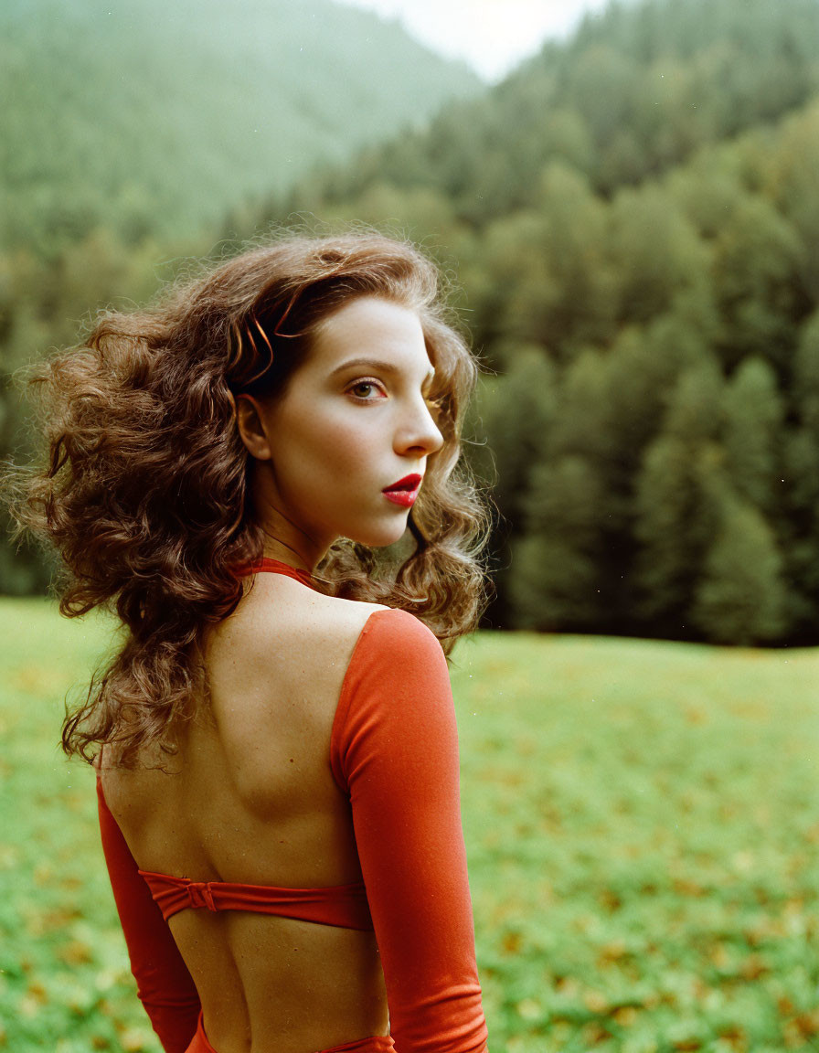 Curly-haired woman in red top gazes over shoulder in lush green meadow.