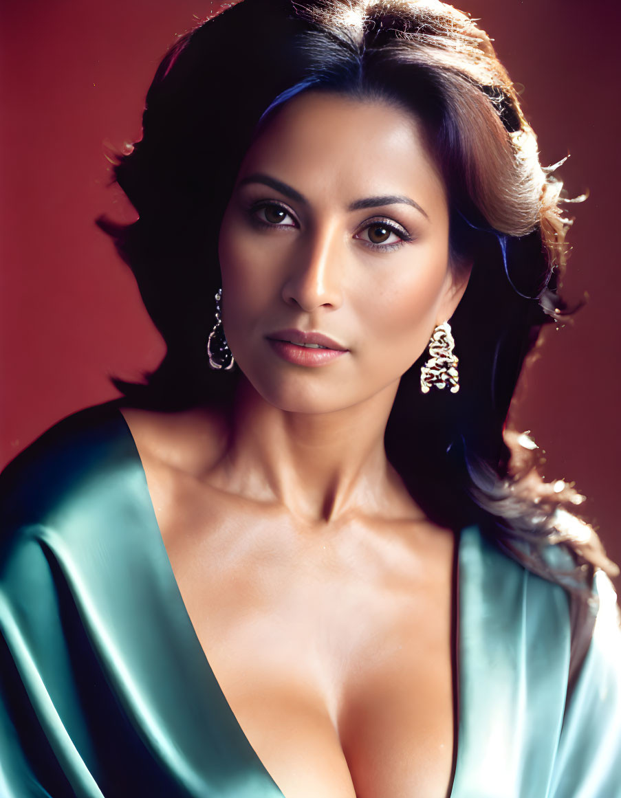 Dark-haired woman in teal satin dress and drop earrings on maroon backdrop