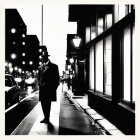 Monochrome image of person in coat and hat on city sidewalk at night