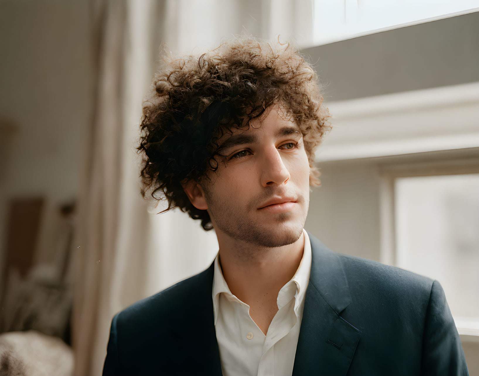 Curly-Haired Young Man in Dark Blazer and White Shirt Gazes Thoughtfully by Bright Window