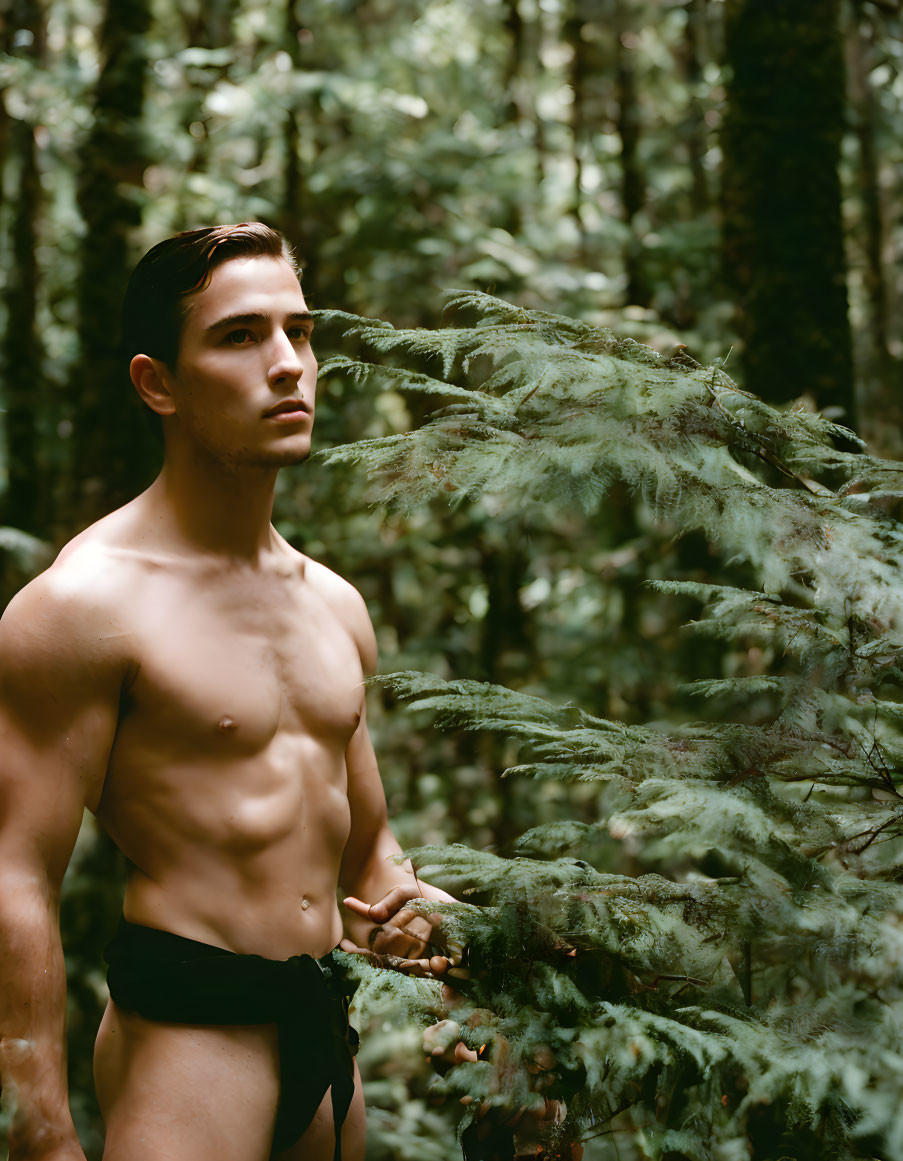 Shirtless man in forest holding fern branch, gazing serenely.