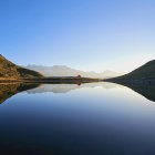 Vibrant surreal landscape: stylized flora, undulating hills, reflective lake, pointed mountains.