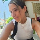 Dark-haired woman in white top and black overalls taking selfie under sunny sky with green umbrellas.