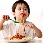 Messy-haired toddler eating happily with red sauce splatter on white bib.